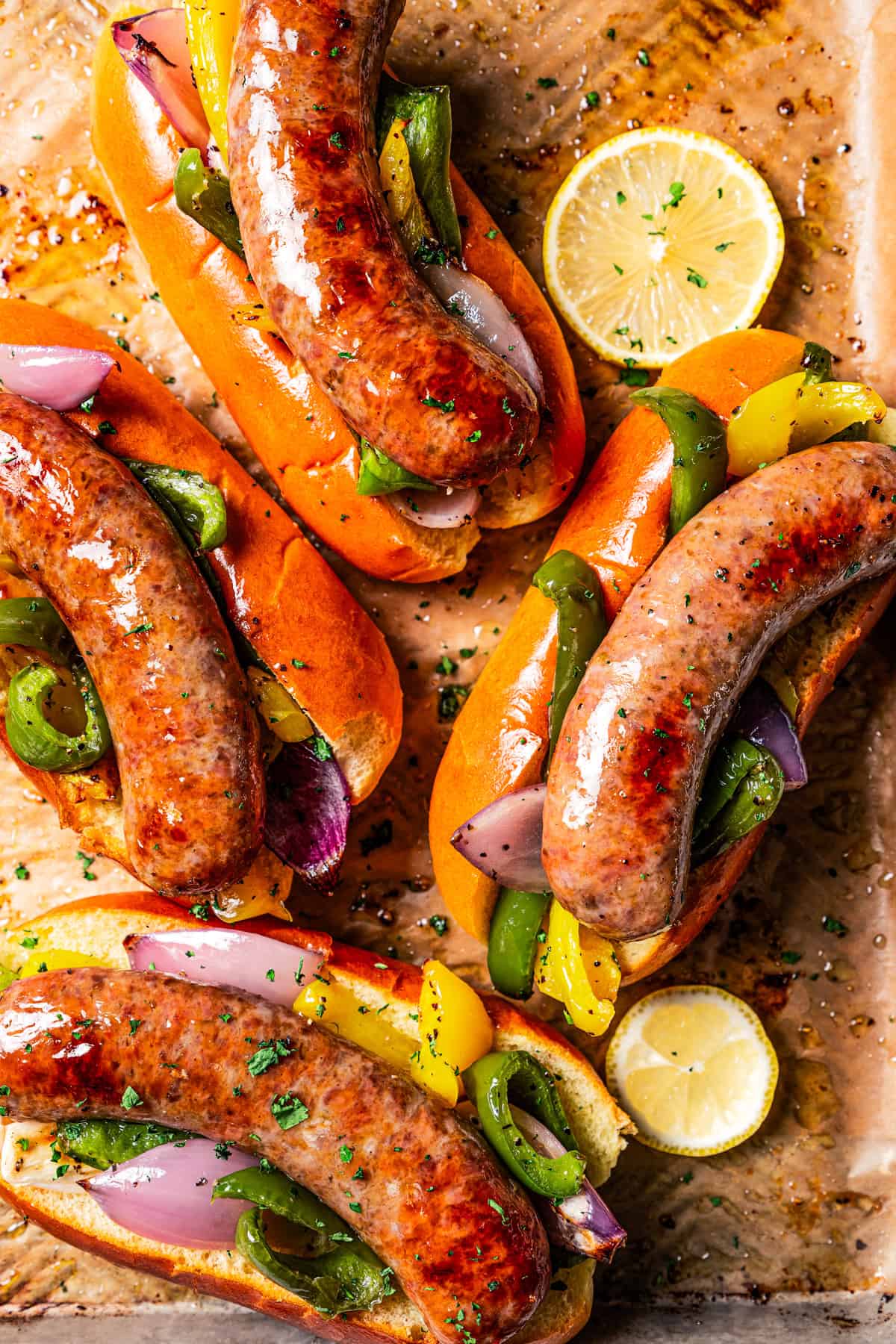 Baked Italian sausage and peppers served on hot dog buns on a baking sheet, next to lemon slices.