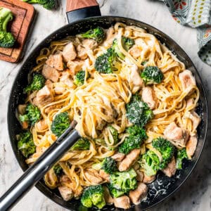 Overhead view of a large serving spoon resting in a pan of chicken fettuccine alfredo with broccoli.