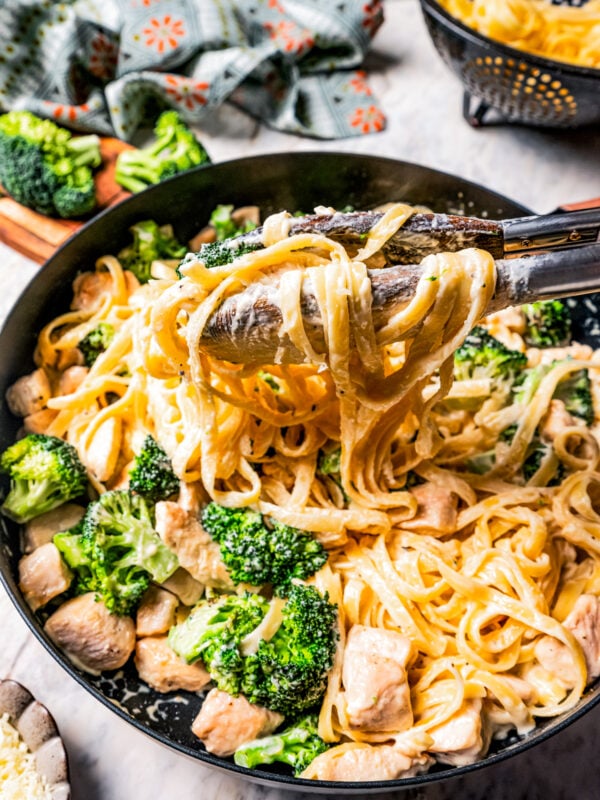 Tongs lifting pasta from a pan of chicken fettuccine alfredo with broccoli.