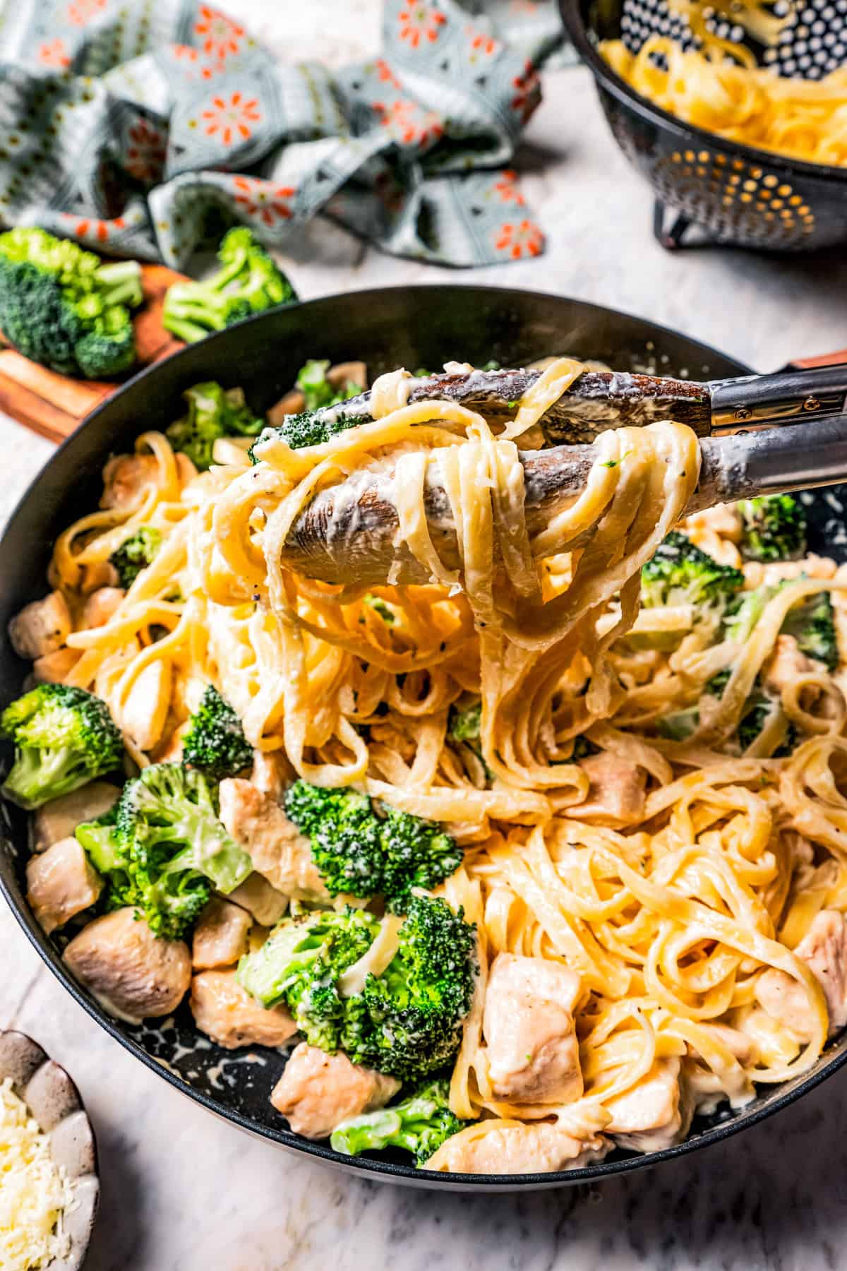 Tongs lifting pasta from a pan of chicken fettuccine alfredo with broccoli.