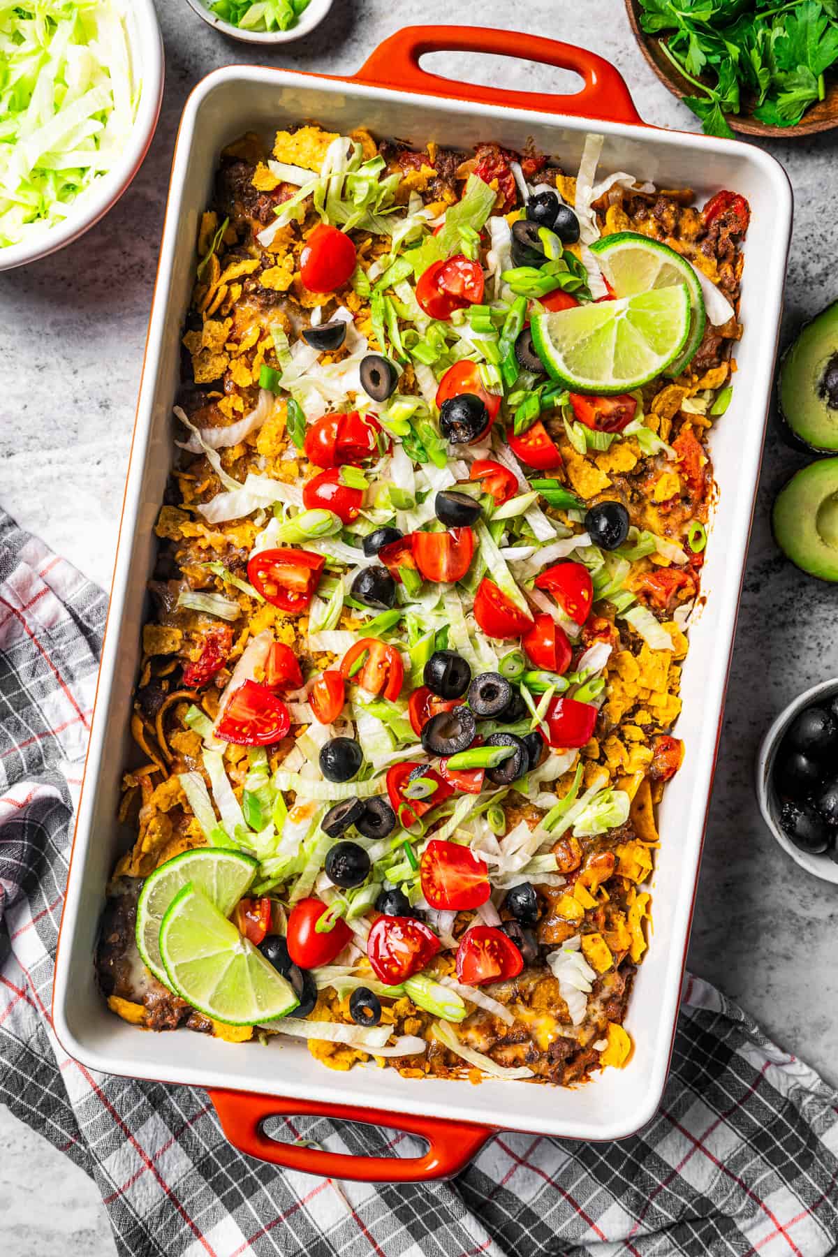 Walking Taco Casserole garnished with shredded lettuce, olives, diced tomatoes, and lime wedges in a baking dish.