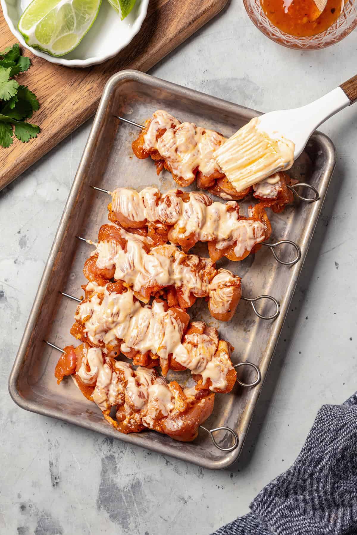 A basting brush brushing seasoned chicken skewers with bang bang sauce on a metal pan.