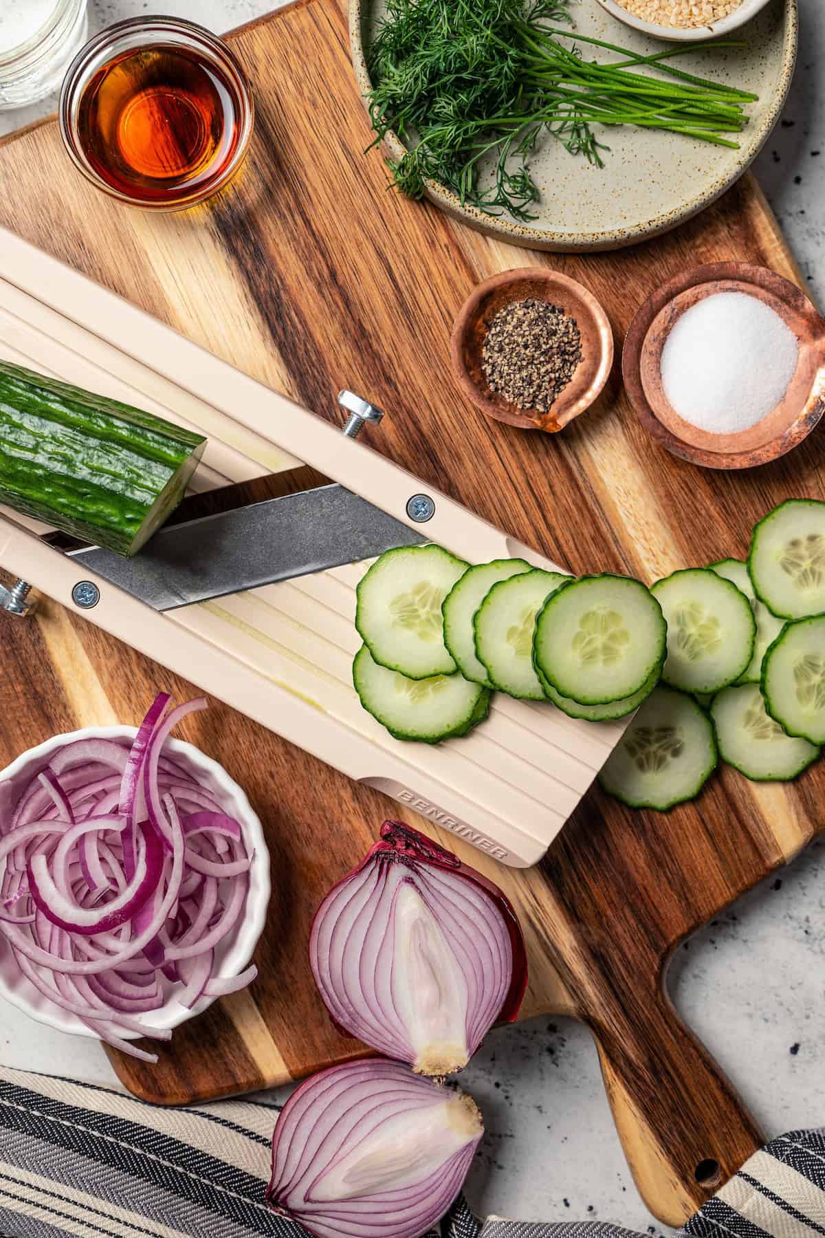 Overhead view of a partially sliced cucumber on a wooden cutting board next to partially sliced onions and other salad ingredients.