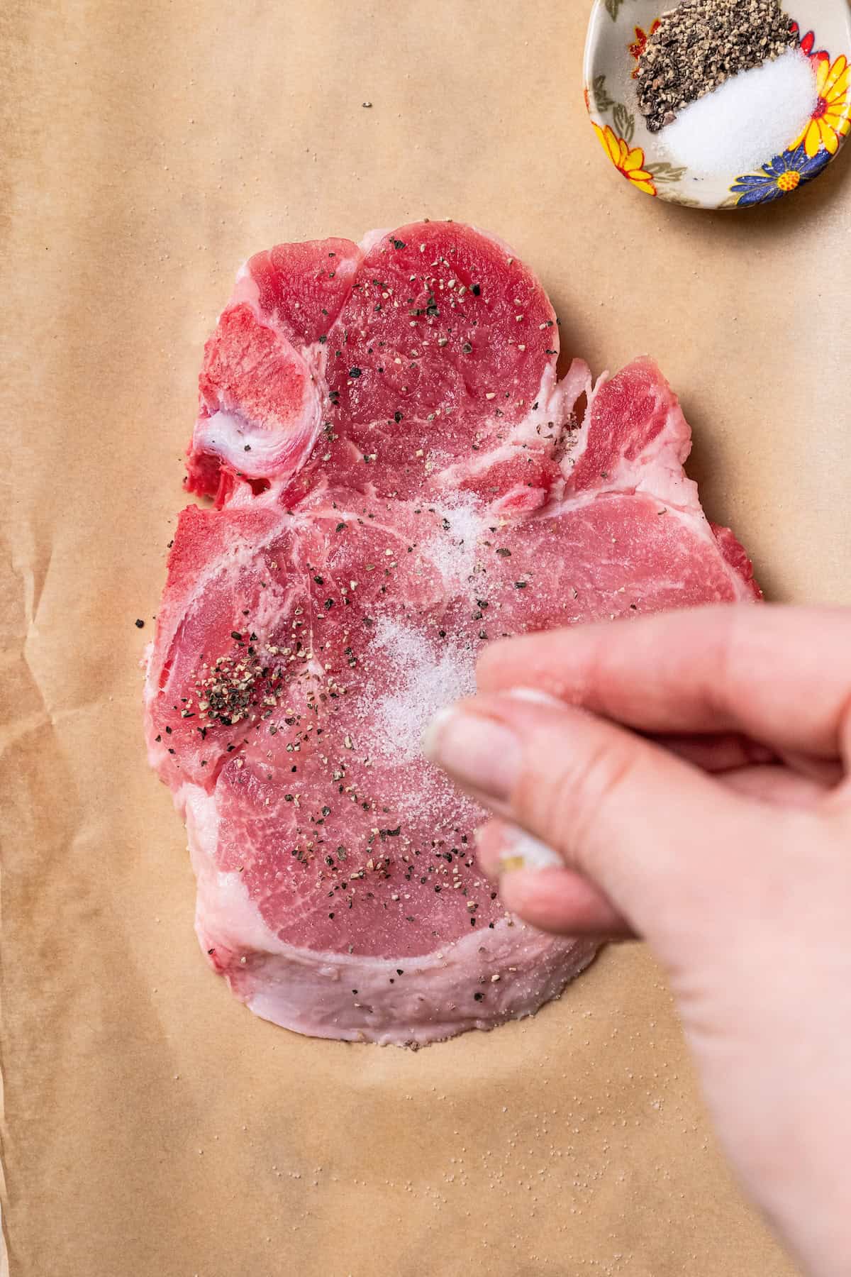 A hand sprinkling salt over a partially seasoned pork steak on a wooden cutting board.