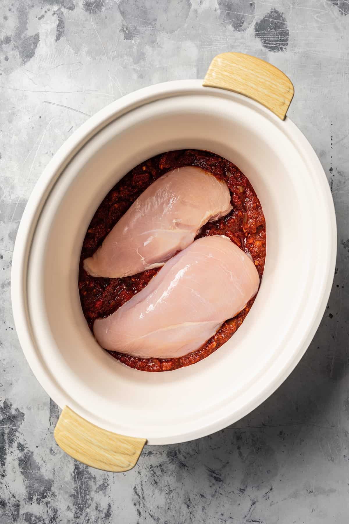 Two uncooked chicken breasts nestled on top of the sauce ingredients inside the crock pot.