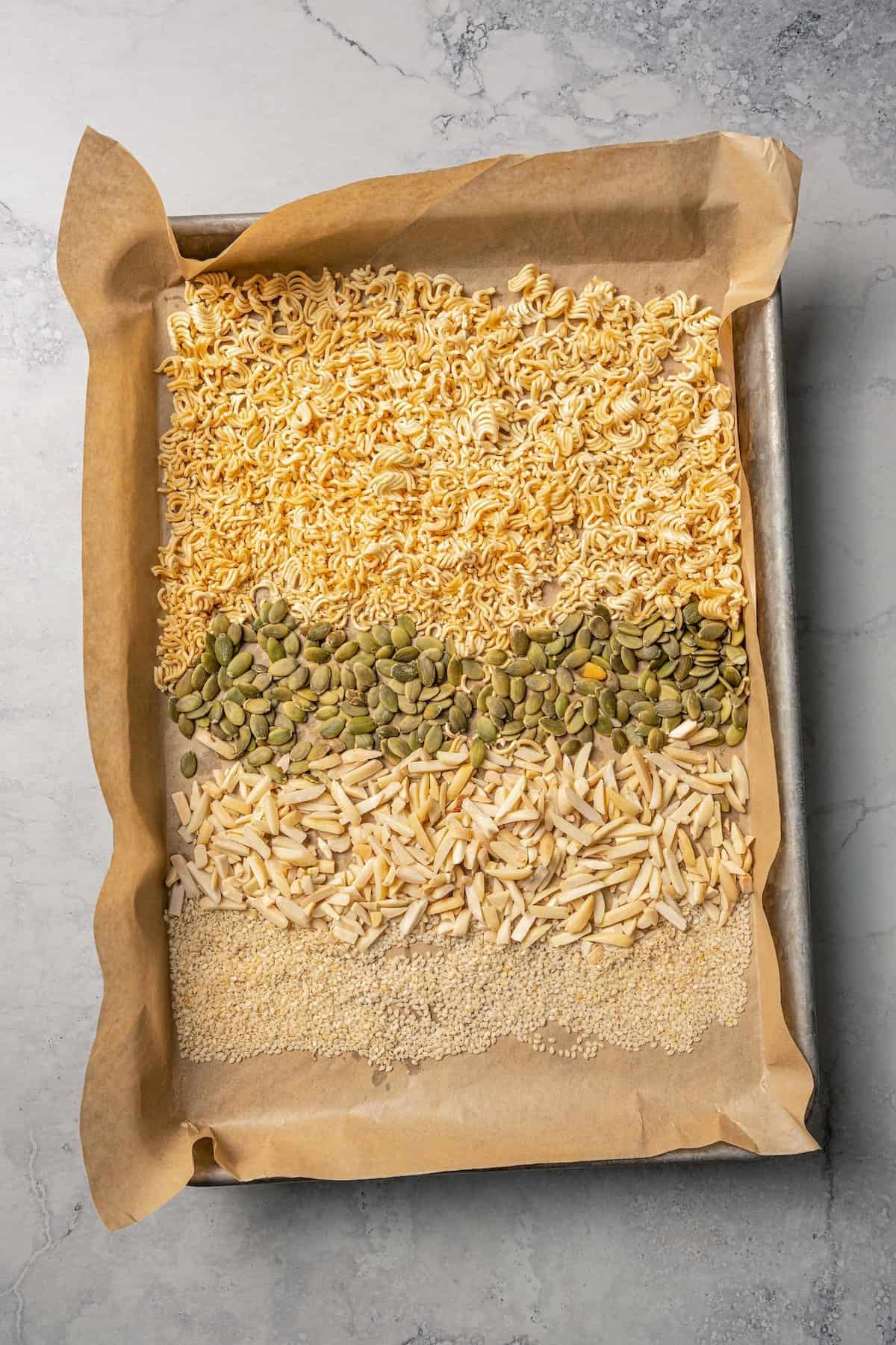 Toasted ramen noodles, slivered almonds, pumpkin, and sesame seeds on a parchment-lined baking sheed.