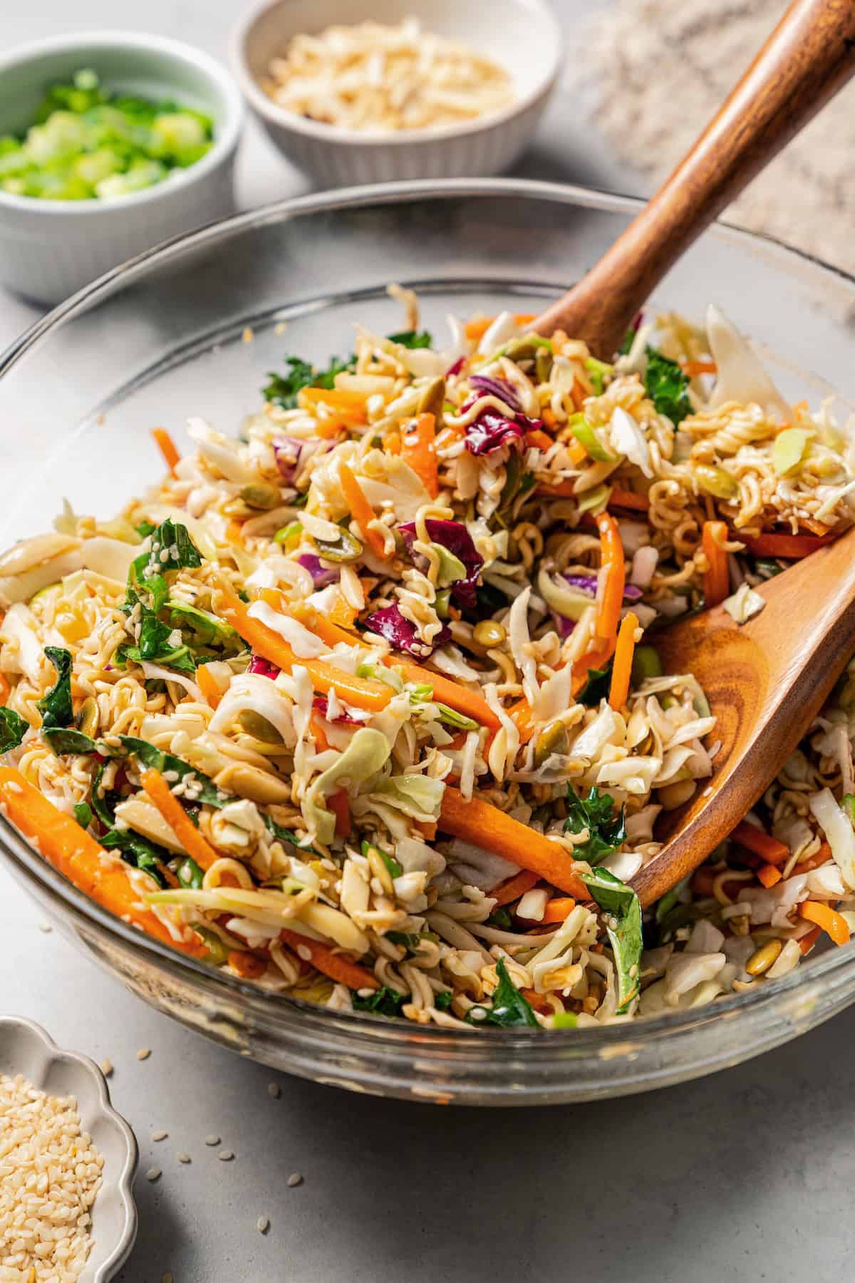 Ramen salad ingredients tossed with dressing in a glass bowl with two wooden salad tongs.