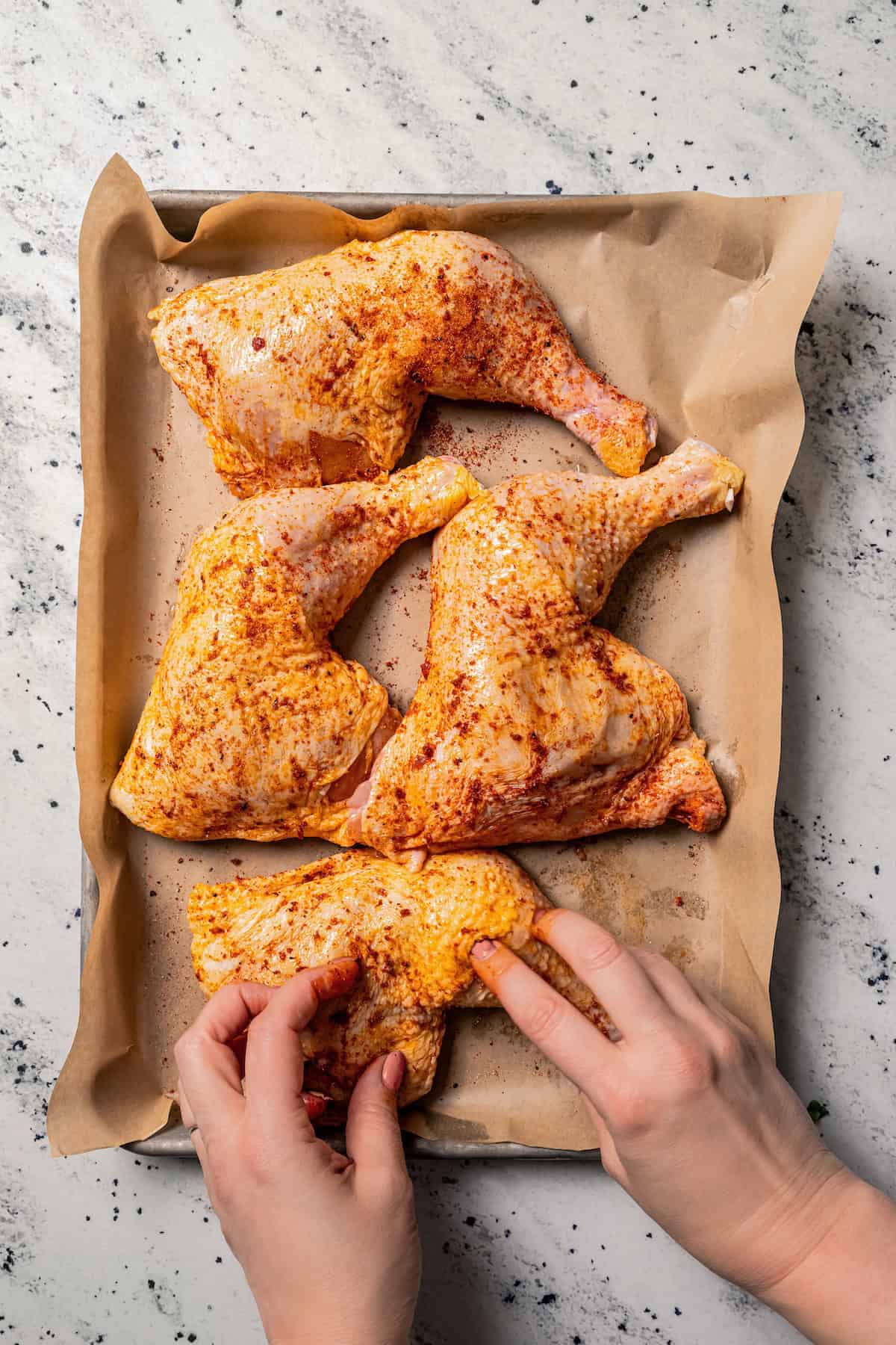 Two hands rubbing seasoning over uncooked chicken leg quarters on a baking pan.