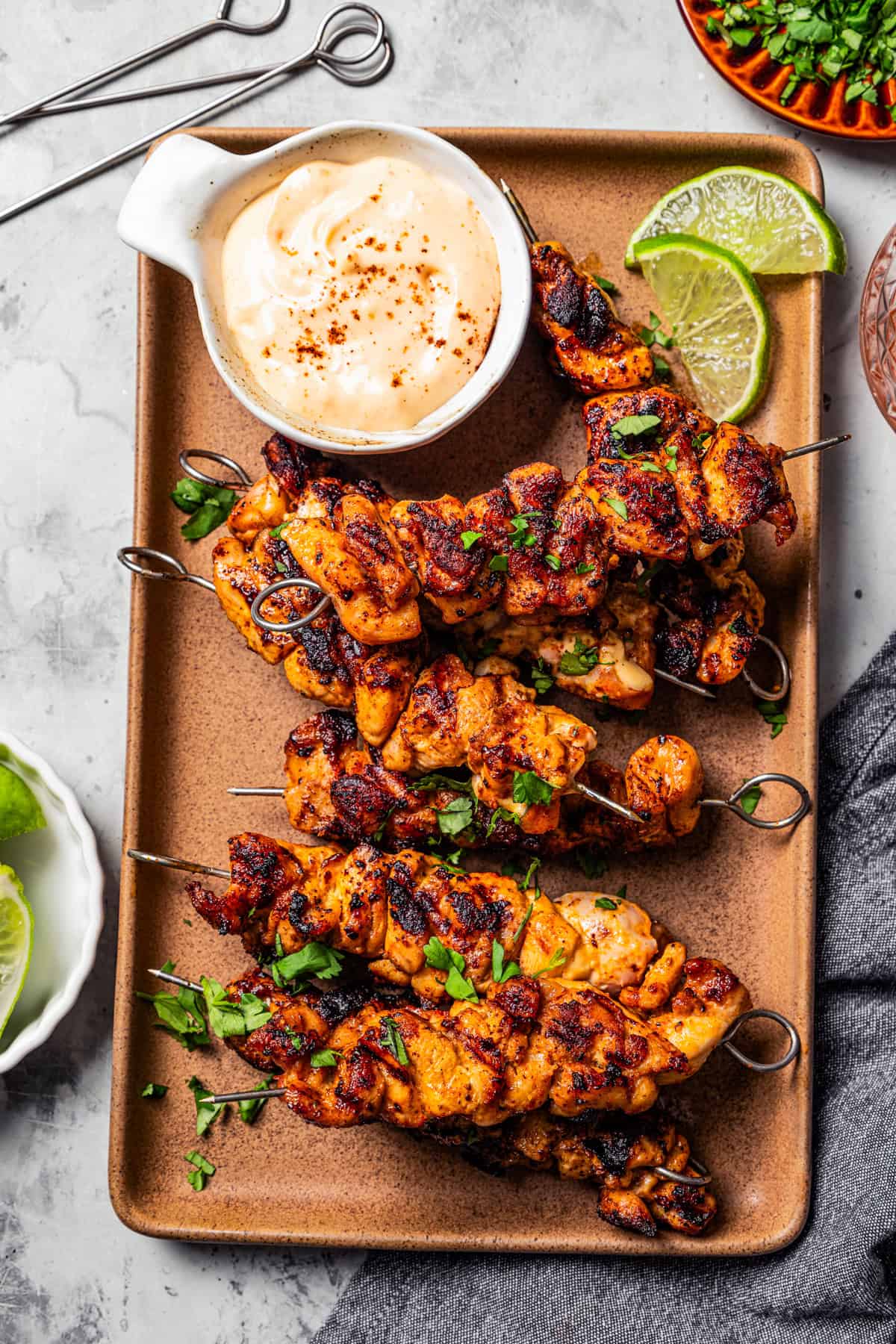 Bang Bang Chicken Skewers A tray of bang bang chicken skewers with a bowl of dipping sauce placed next to the skewers, and garnished with cilantro and lime wedges.