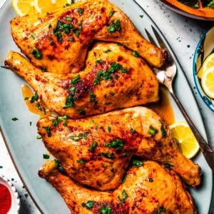 chicken leg quarters served on a large platter garnished with lemon slices, with a set of serving utensils.