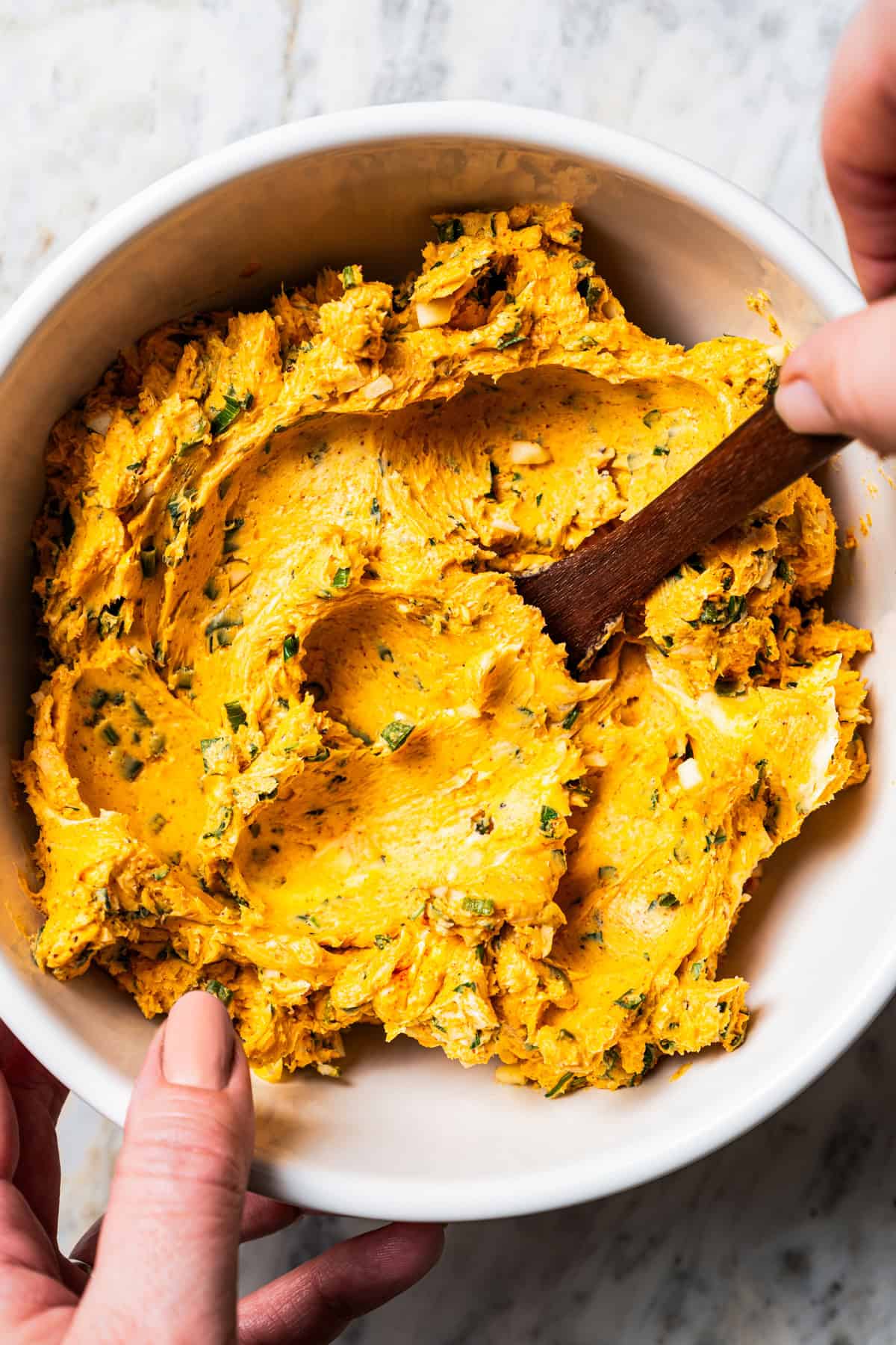 A hand holding a bowl of cowboy butter while the other hand stirs with a wooden spatula.