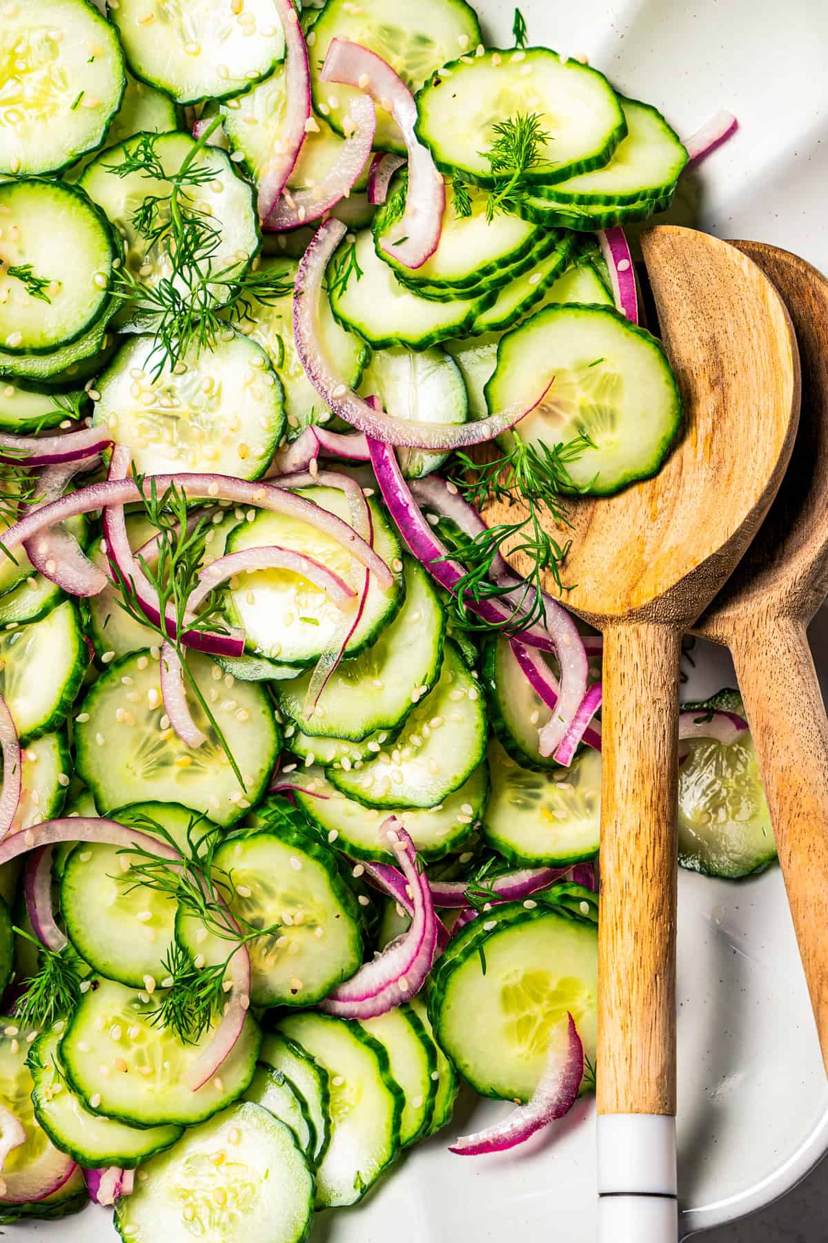 Overhead view of cucumber and onion salad in a large scalloped salad bowl with two wooden salad tongs.