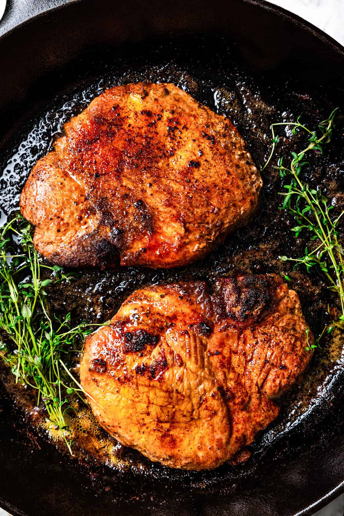 Two seared pork steaks in a cast iron skillet with fresh thyme sprigs.