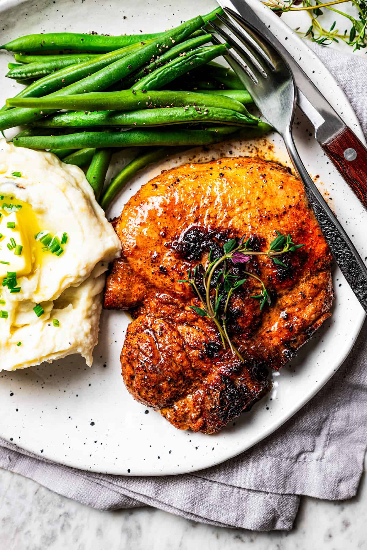 Pork steak garnished with thyme sprigs next to a serving of mashed potatoes and green beans on a plate, with a fork and knife.