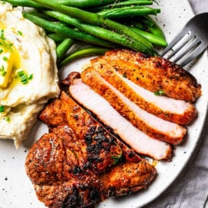 Overhead view of a fork resting next to a sliced pork steak with a side of mashed potatoes and green beans on a plate.