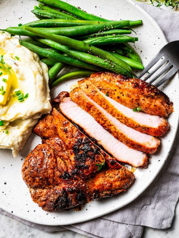 Overhead view of a fork resting next to a sliced pork steak with a side of mashed potatoes and green beans on a plate.