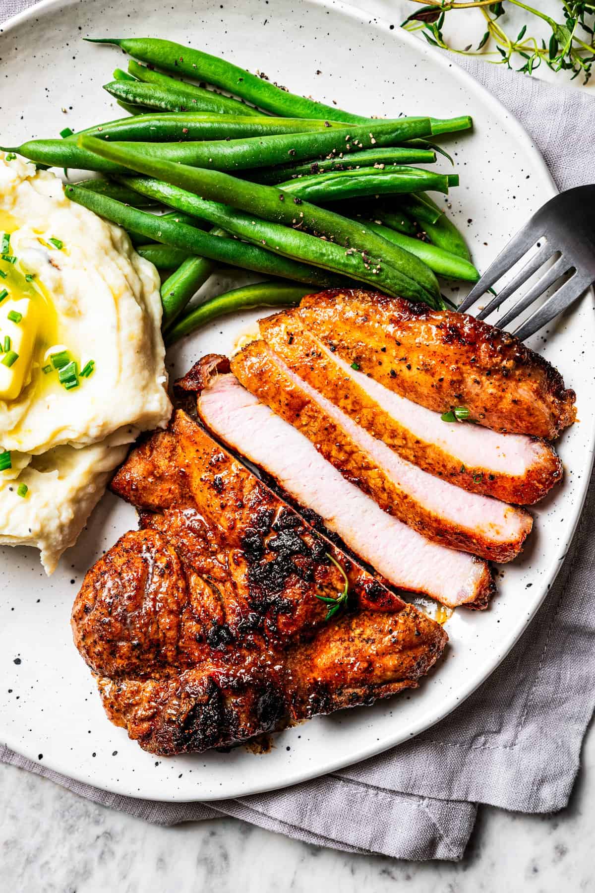 Overhead view of a fork resting next to a sliced pork steak with a side of mashed potatoes and green beans on a plate.