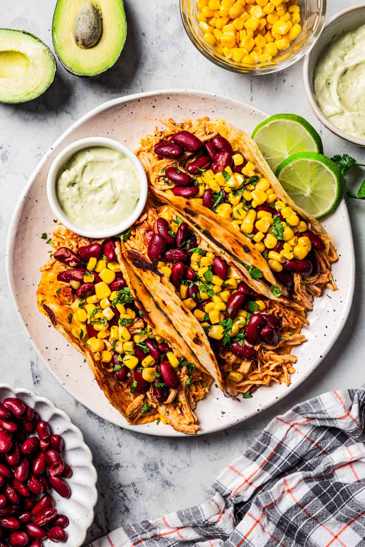 Overhead view of three pulled chicken tacos topped with corn and beans next to a bowl of dipping sauce on a plate.