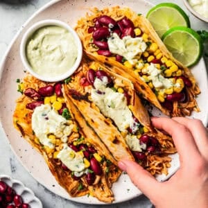 Overhead view of a hand lifting a pulled chicken taco topped with avocado sauce from a trio of tacos served on a plate.