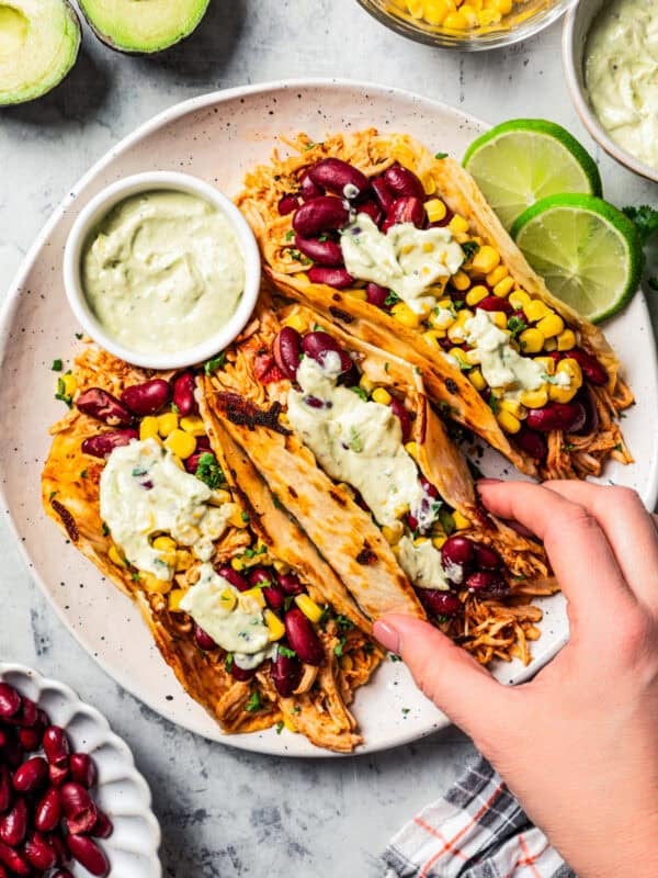 Overhead view of a hand lifting a pulled chicken taco topped with avocado sauce from a trio of tacos served on a plate.