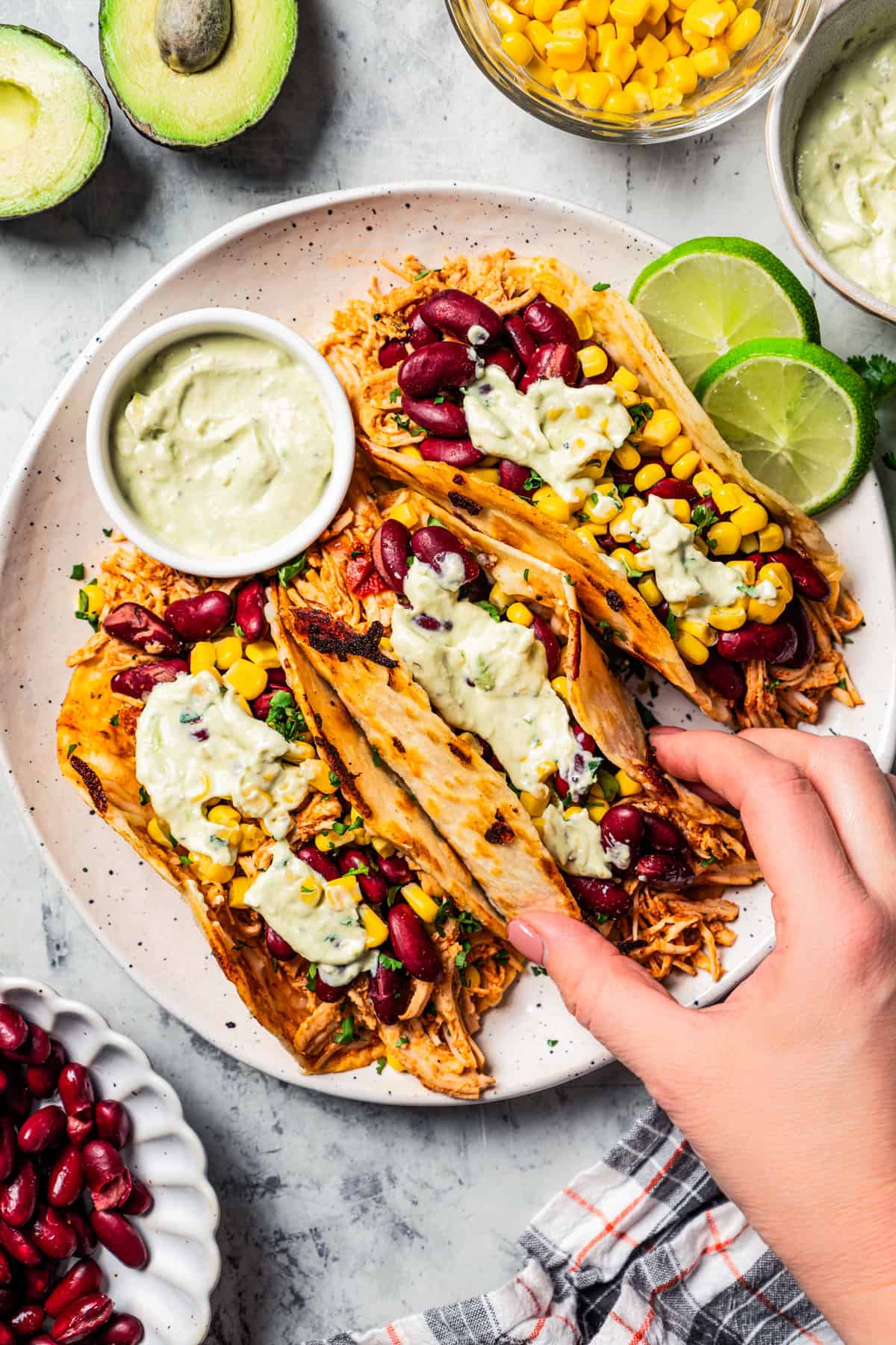 Overhead view of a hand lifting a pulled chicken taco topped with avocado sauce from a trio of tacos served on a plate.