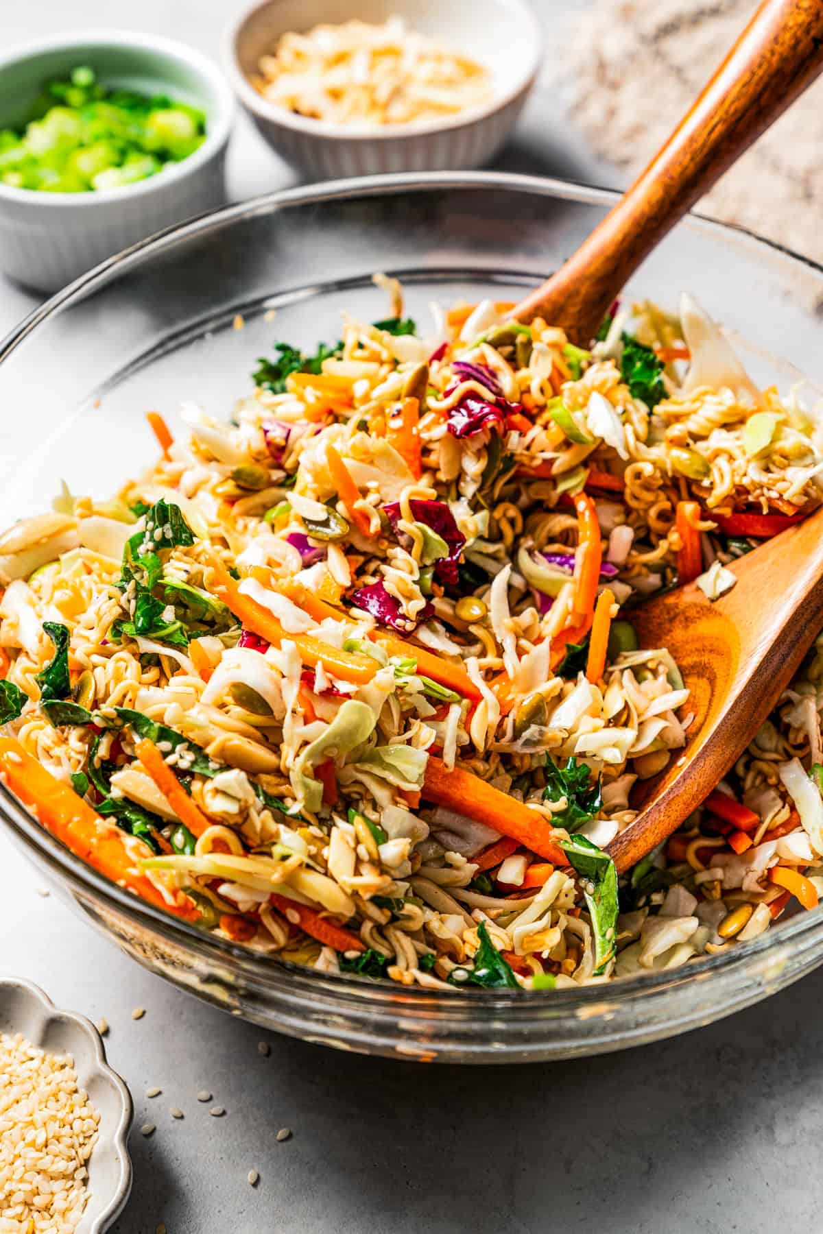 Ramen salad ingredients tossed with dressing in a glass bowl with two wooden salad tongs.