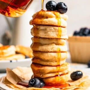 Pouring maple syrup over a stack of silver dollar pancakes.