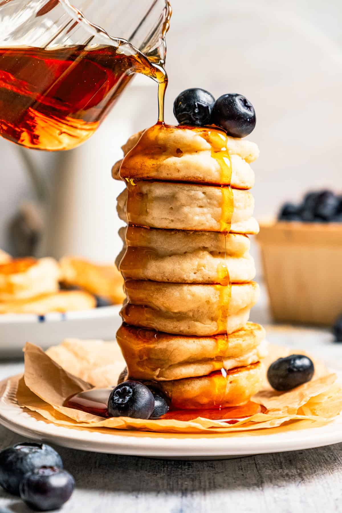 Pouring maple syrup over a stack of silver dollar pancakes.