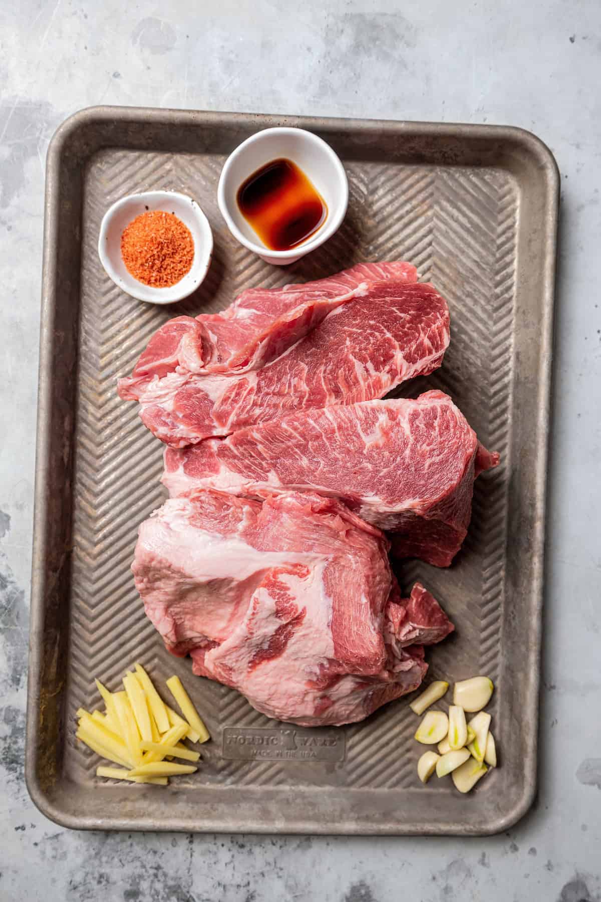 Kalua pork (Hawaiian pulled pork) is made from seasoned pork shoulder rubbed with sea salt and slow-roasted until it's fall-apart tender. Pork shoulder cut into three pieces on a baking sheet next to small bowls of seasoning.