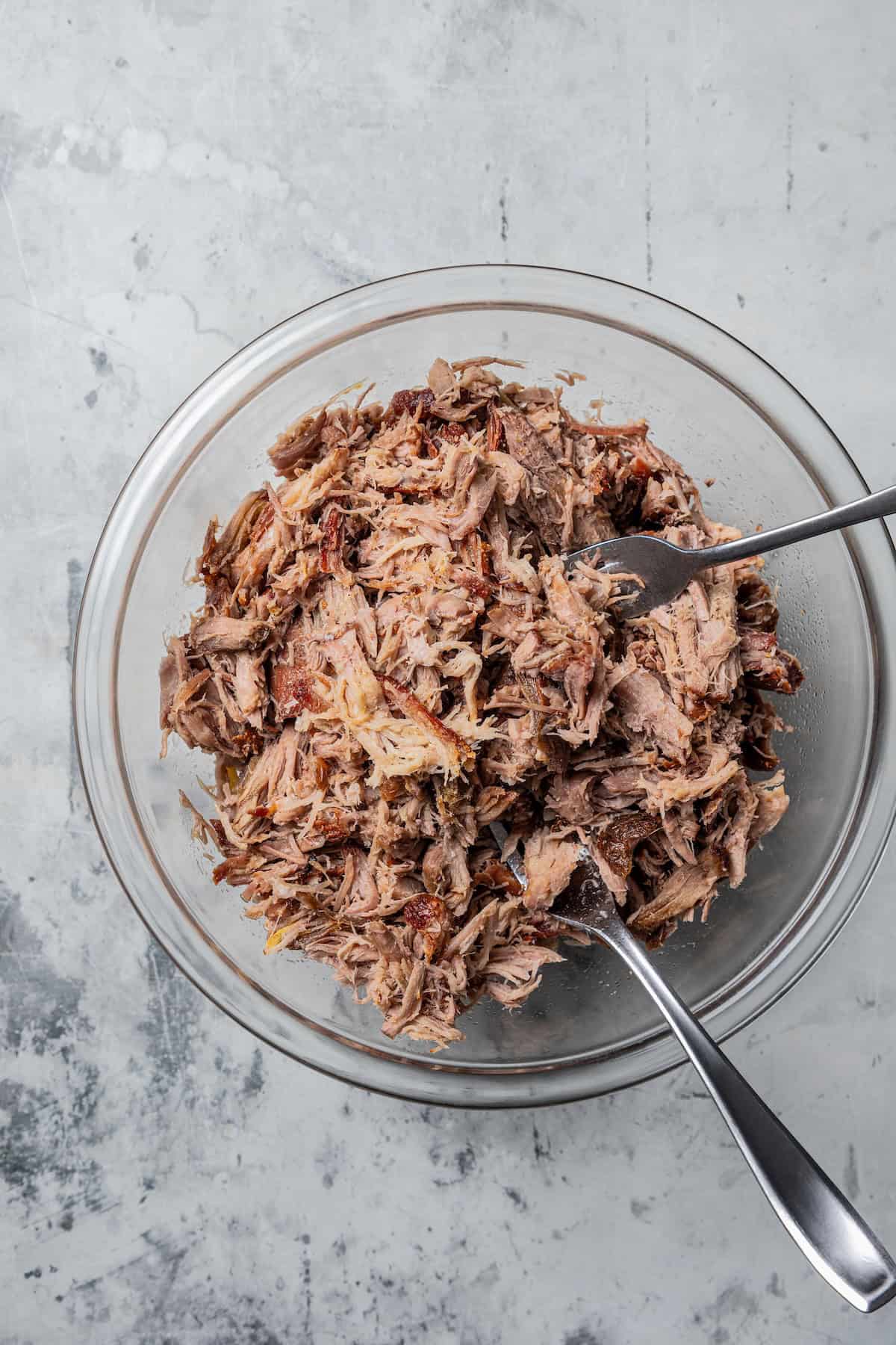 Kalua pork (Hawaiian pulled pork) is made from seasoned pork shoulder rubbed with sea salt and slow-roasted until it's fall-apart tender. Shredded pork in a glass bowl with two forks.