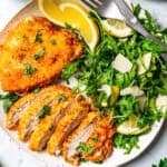 Sliced chicken schnitzel next to a whole schnitzel on a plate, with a side of green salad and lemon wedges.