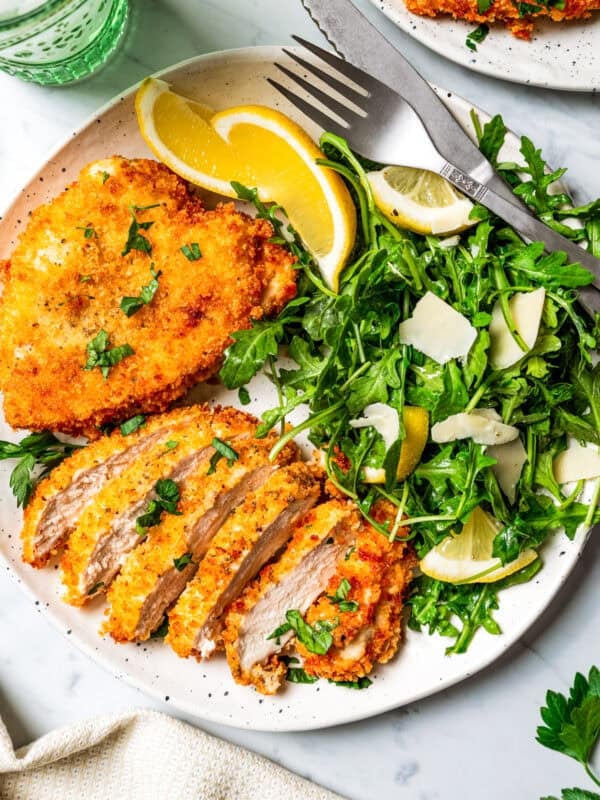 Sliced chicken schnitzel next to a whole schnitzel on a plate, with a side of green salad and lemon wedges.