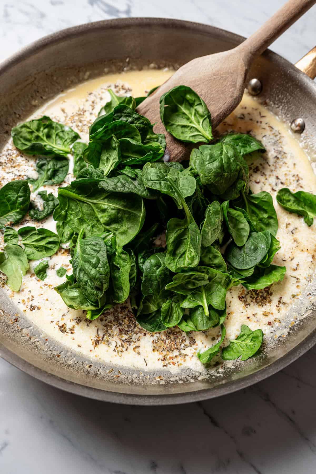 Creamy chicken Florentine is a one-pan dinner with golden-fried chicken smothered in a garlicky spinach cream sauce. Make it in 30 minutes! Baby spinach added to cream sauce in a skillet with a wooden spoon.
