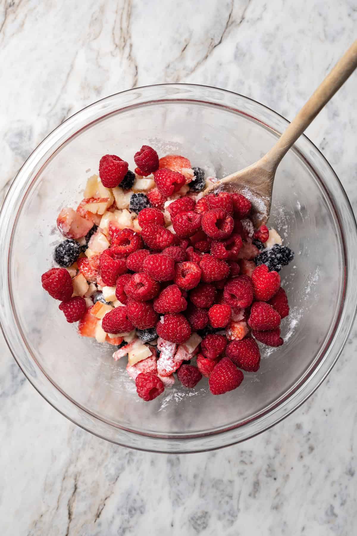 This easy mixed berry pie recipe combines three types of juicy berries with tender apples inside a light and flaky homemade pie crust. Apples and berries combined in a bowl with a wooden spoon.
