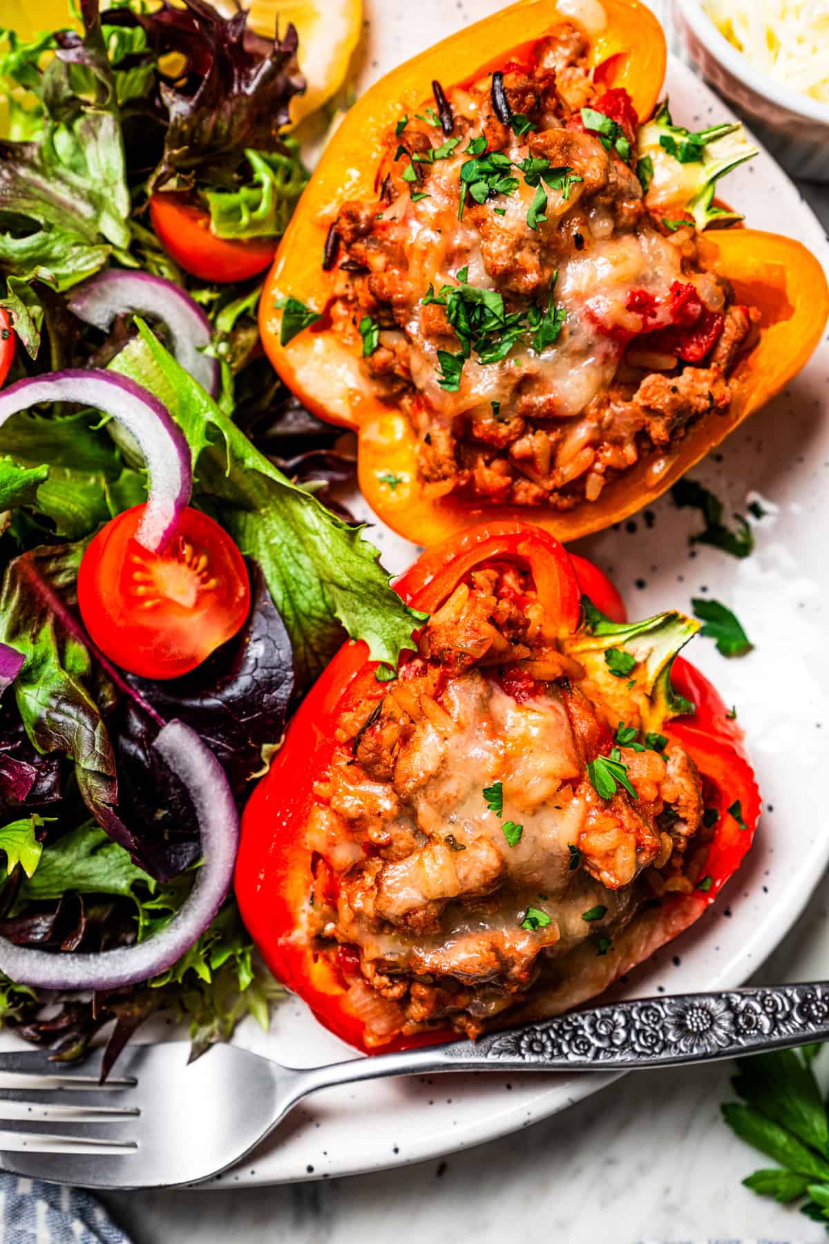 Esses pimentões recheados de peru moídos clássicos estão repletos de carne moída temperada, arroz selvagem e tomate suculento, coberto com queijo derretido e assado. Dois pimentões recheados de peru moídos ao lado de um lado de salada verde em um prato. Foto de dietood.com