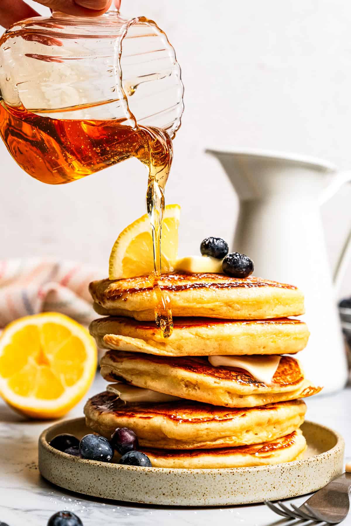 Maple syrup is being poured from a glass jug over a stack of lemon ricotta pancakes on a stoneware plate.