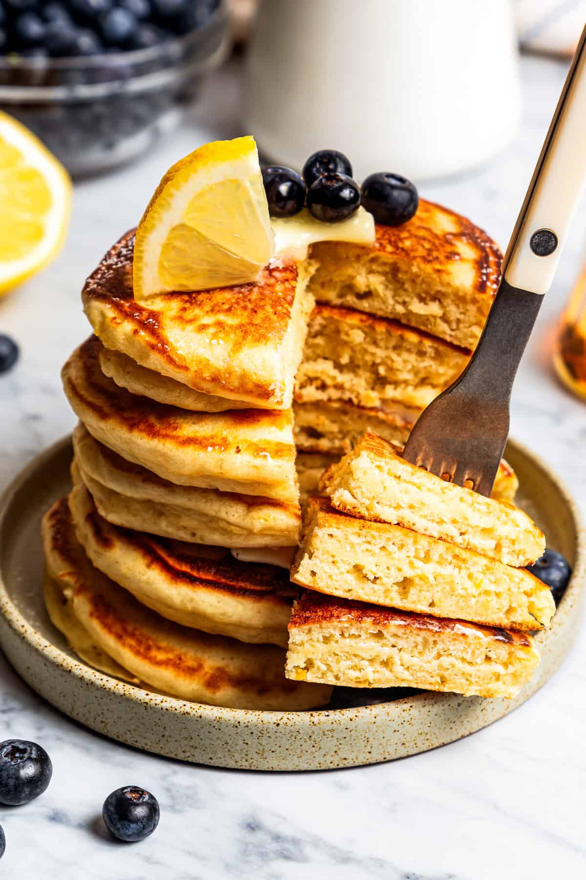 A fork stuck into a wedge of stacked lemon ricotta pancakes cut from the rest of the stack on a stoneware plate.