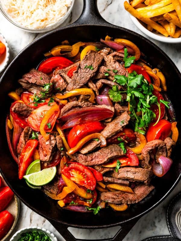 Overhead view of lomo saltado in a cast-iron skillet next to bowls of rice and fries.