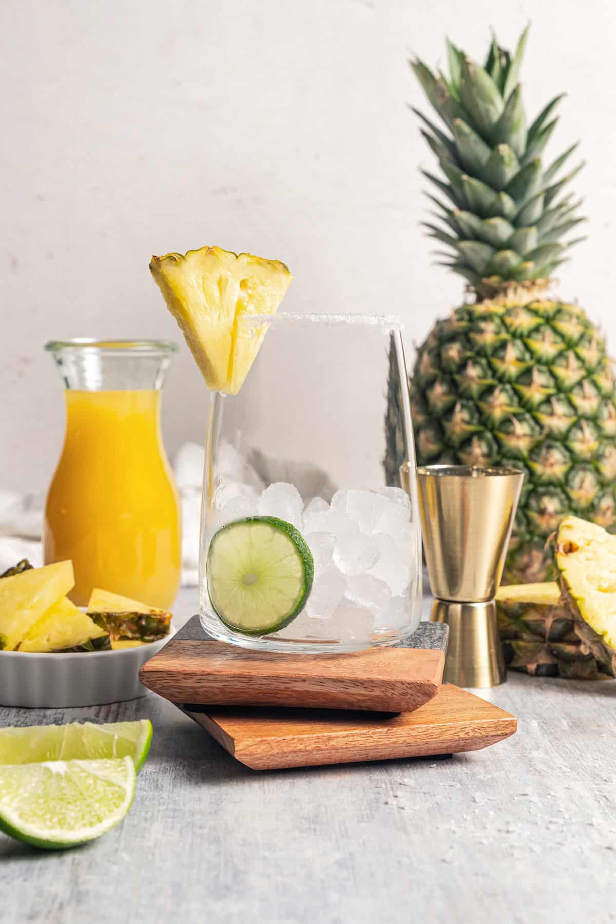 This easy pineapple margarita is a juicy blend of tequila blanco, fresh pineapple juice, and lime, shaken over ice. Don’t forget a salt rim! A glass filled with ice, rimmed with salt, and garnished with a pineapple wedge and lime slices wth pineapple margarita ingredients in the background.