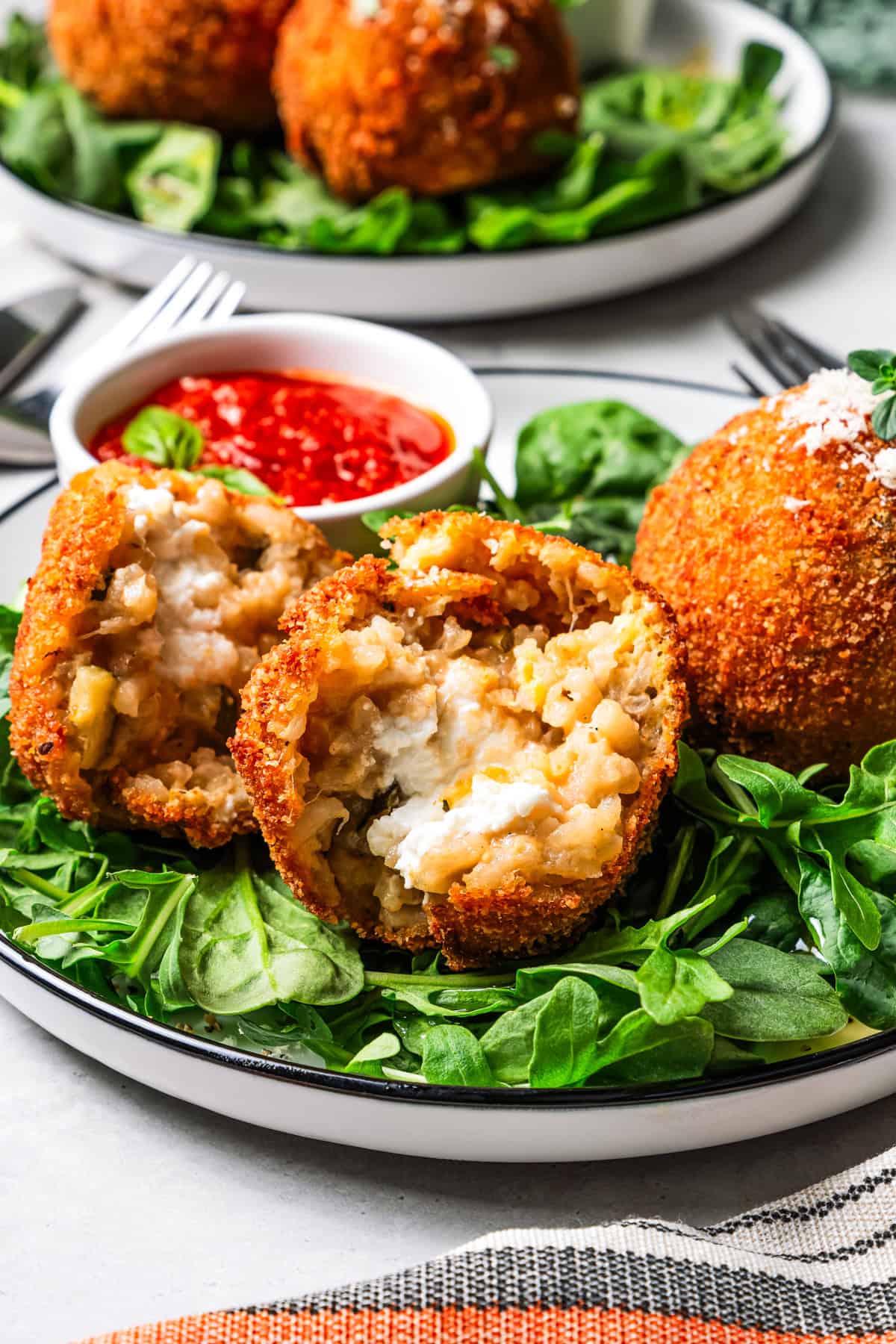 This easy homemade arancini recipe makes crispy, cheesy, golden-fried Italian rice balls filled with melty pockets of tangy goat cheese. An arancini ball broken in half and served over a bed of greens on a plate.