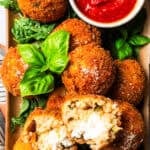 Arancini ball broken in half on top of a pile of arancini on a platter, next to a ramekin of marinara sauce.