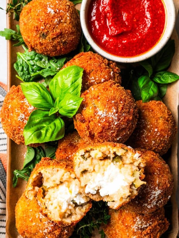 Arancini ball broken in half on top of a pile of arancini on a platter, next to a ramekin of marinara sauce.