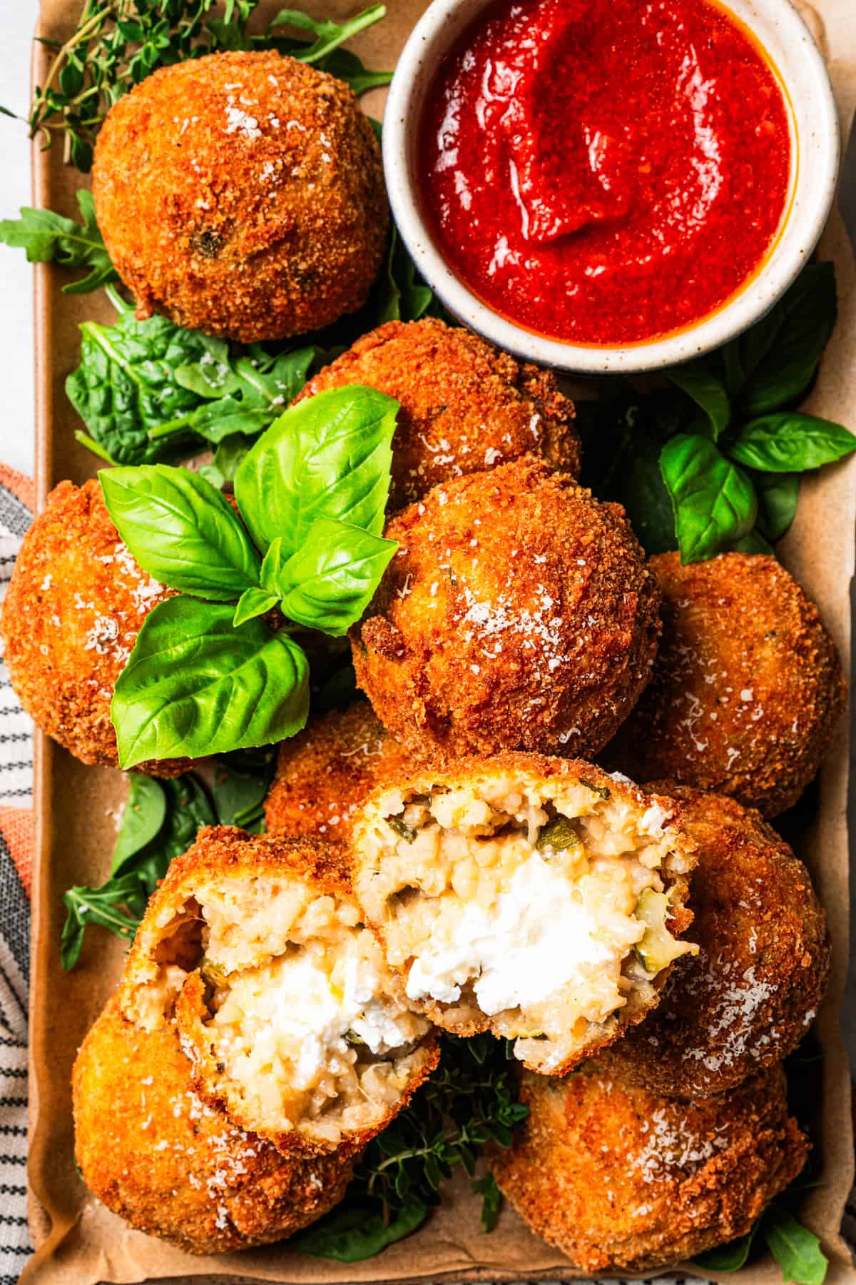 This easy homemade arancini recipe makes crispy, cheesy, golden-fried Italian rice balls filled with melty pockets of tangy goat cheese. Arancini ball broken in half on top of a pile of arancini on a platter, next to a ramekin of marinara sauce.