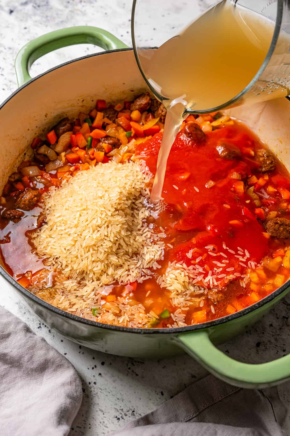 Classic arroz con pollo is a Latin favorite with juicy, seasoned chicken and rice brimming with smoky, savory flavor. Make it in one pot! Broth is being poured over the rice ingredients in a large pot.
