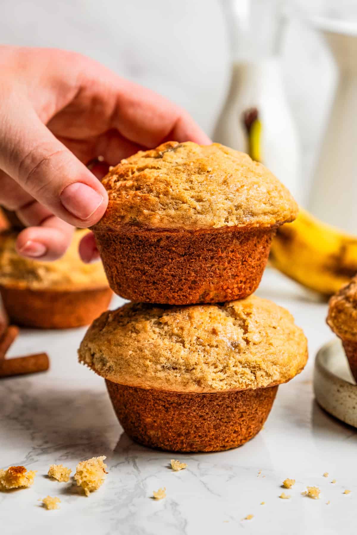 These soft, buttery banana bread muffins are easy to make with maple syrup, brown sugar, and cinnamon. It's impossible to eat just one! A hand picking up a banana bread muffin from a stack of two muffins.
