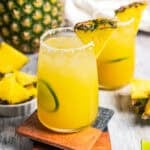 A pineapple margarita in a salt-rimmed glass garnished with a pineapple wedge and lime slice, with a second margarita in the background. Photo by diethood.