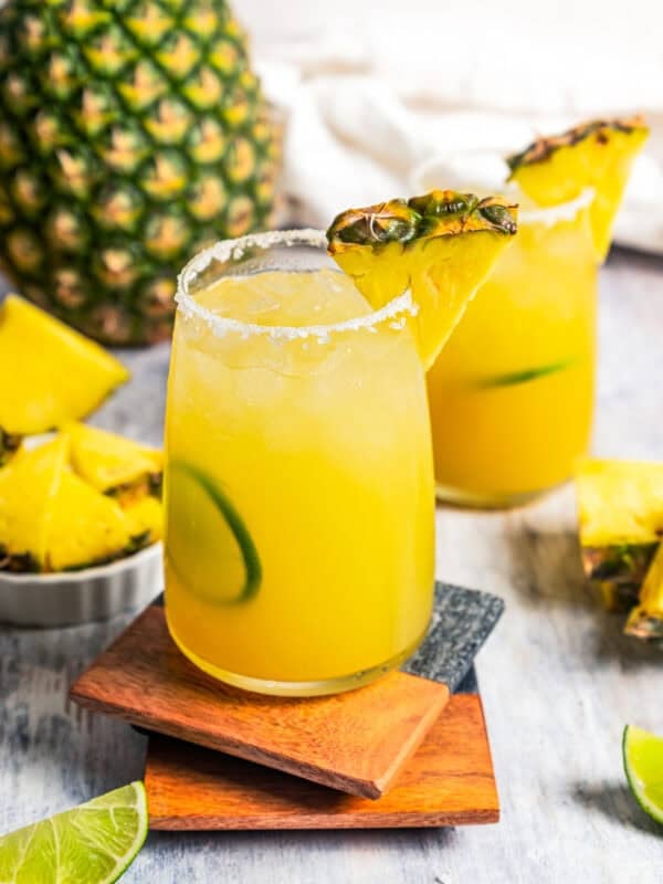 A pineapple margarita in a salt-rimmed glass garnished with a pineapple wedge and lime slice, with a second margarita in the background. Photo by diethood.