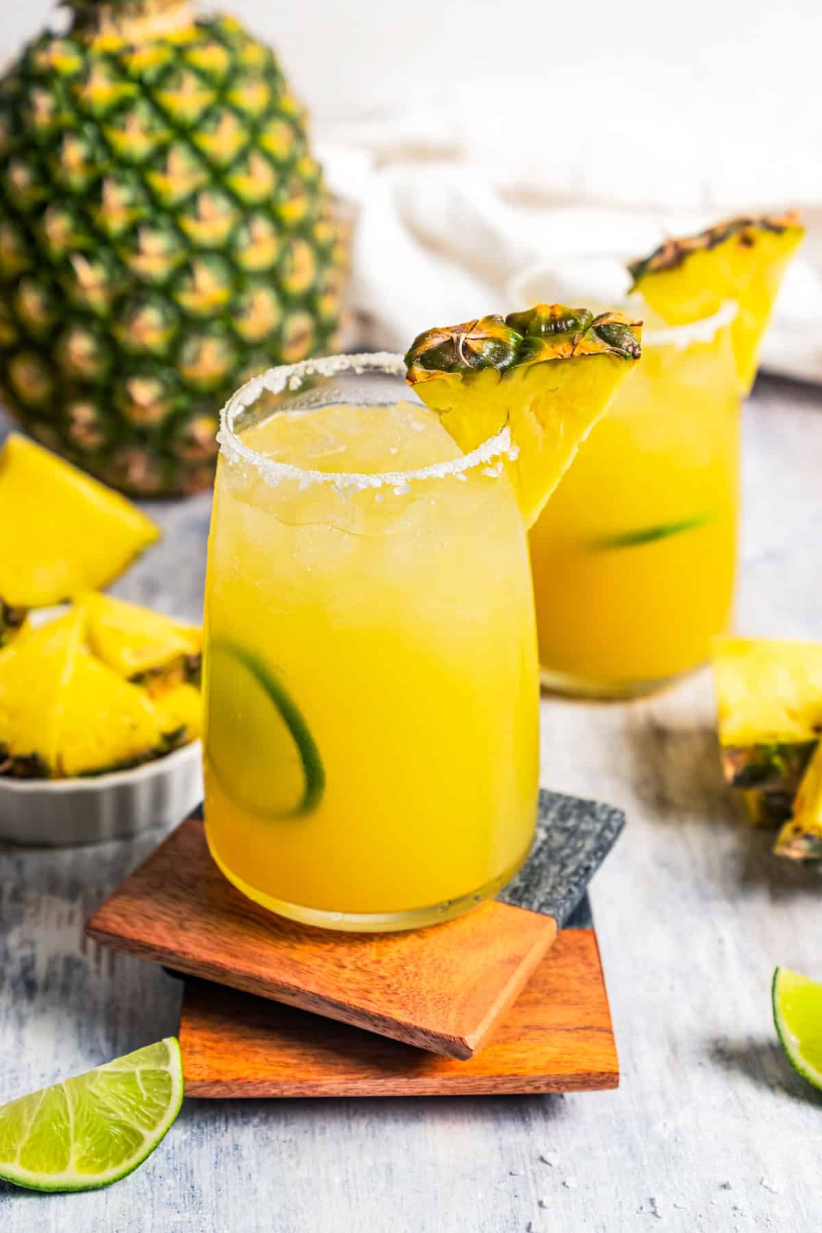 This easy pineapple margarita is a juicy blend of tequila blanco, fresh pineapple juice, and lime, shaken over ice. Don’t forget a salt rim! A pineapple margarita in a salt-rimmed glass garnished with a pineapple wedge and lime slice, with a second margarita in the background. Photo by diethood.