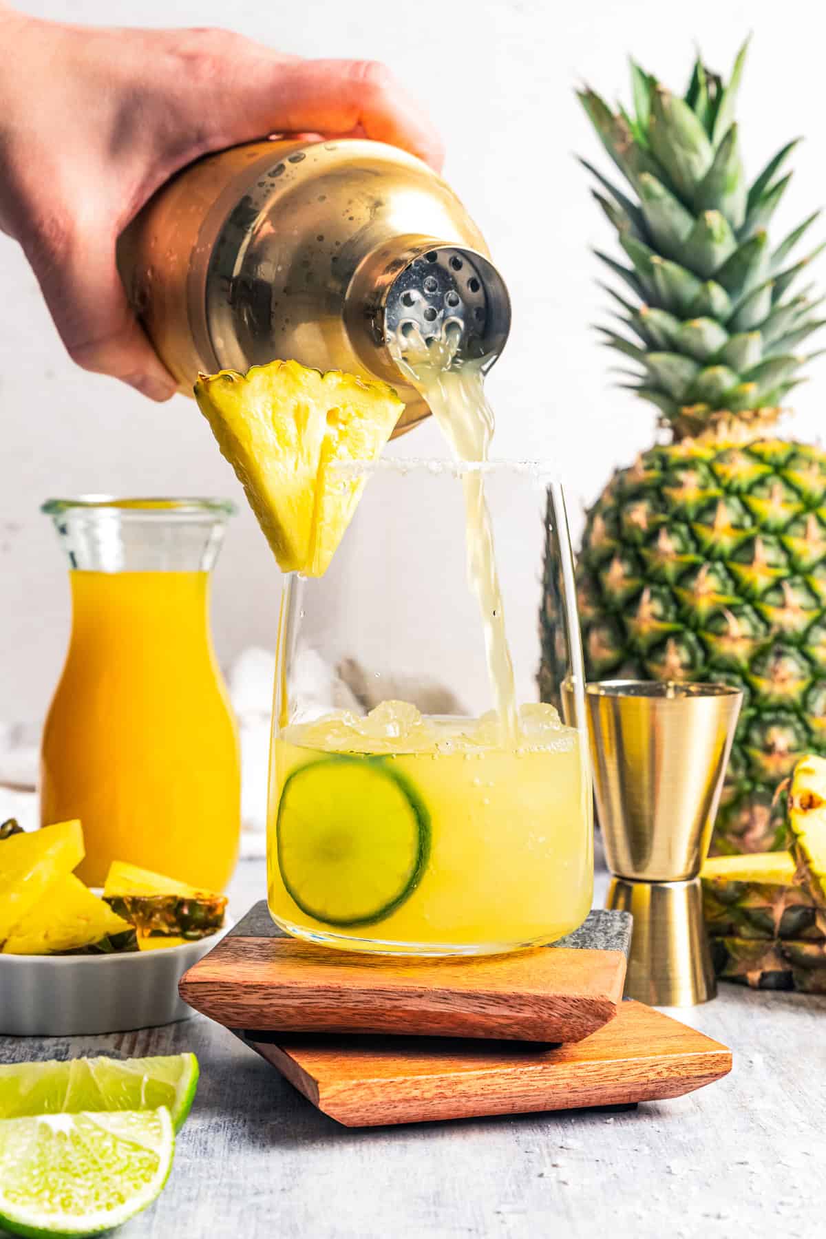 This easy pineapple margarita is a juicy blend of tequila blanco, fresh pineapple juice, and lime, shaken over ice. Don’t forget a salt rim! Pouring a margarita from a cocktail shaker into a glass filled with ice, rimmed with salt, and garnished with a pineapple wedge and lime slices.