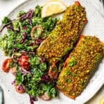 Two pistachio crusted salmon filets with a side of kale salad on a plate.